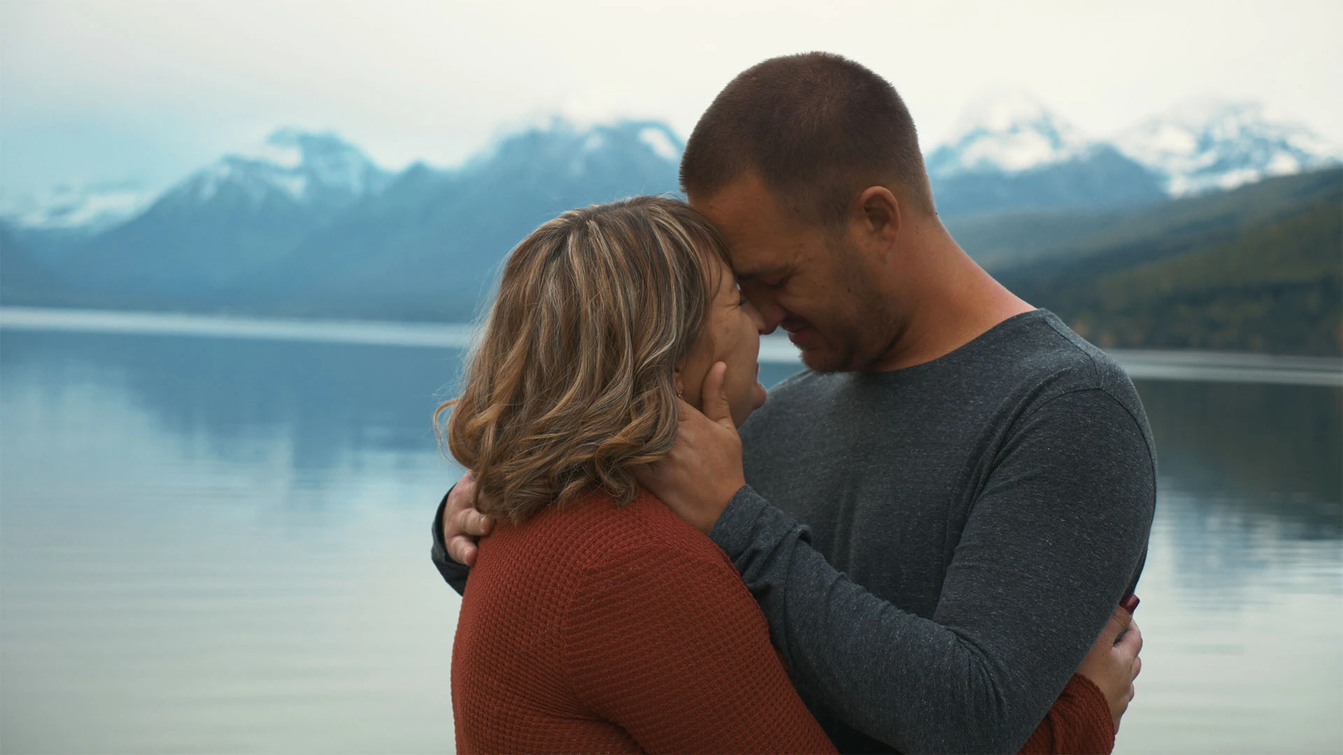 Thomas and Sadie forehead to forehead beside a still Montana lake below snowcapped peaks